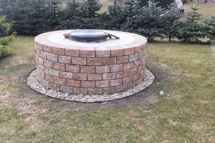 Okrągła studnia głębinowa z cegły, z metalową pokrywą. Wokół żwir i trawnik. Sprawdź studnia głębinowa koszt.